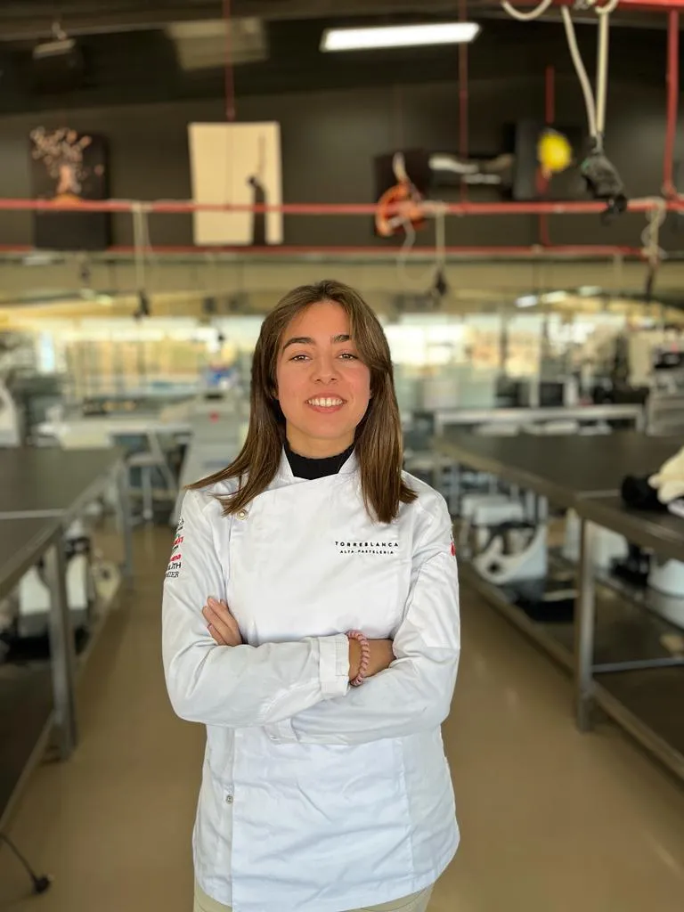 Carmen posando sonriente con chaquetilla blanca de pastelera en el obrador de la escuela Torre Blanca.