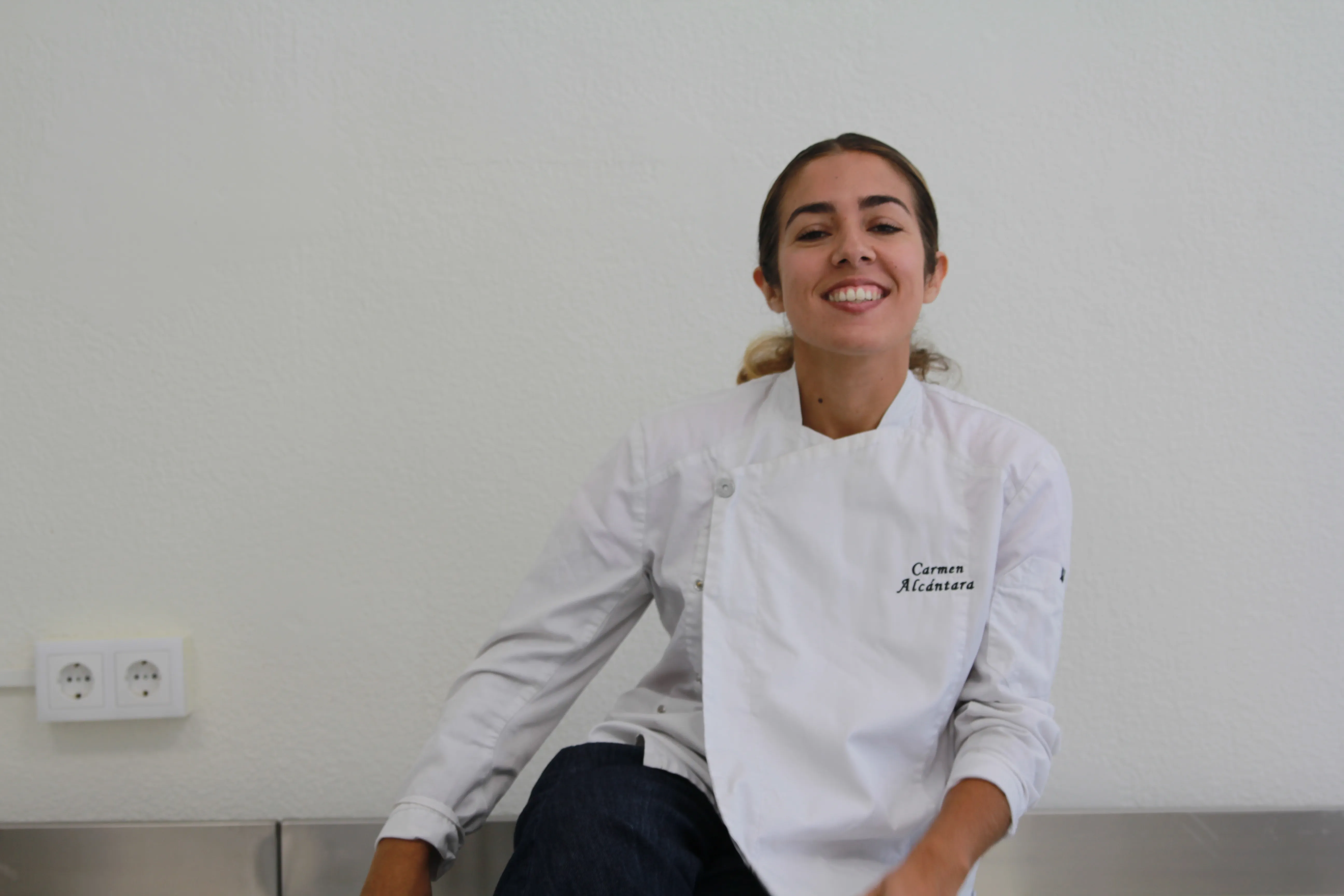 Sonriendo con chaquetilla blanca de pastelera, sentada en una encimera de acero contra una pared blanca.