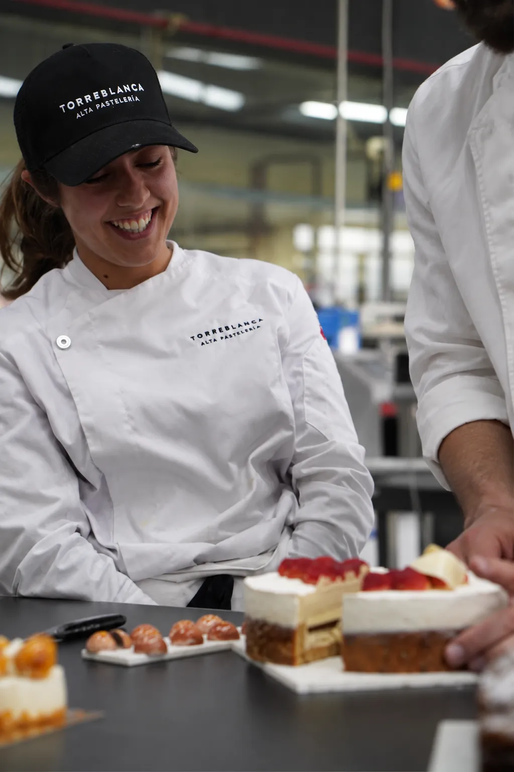 Sonriendo con gorra y chaquetilla de Torreblanca ante una mesa de pasteles con fresas, en el obrador.