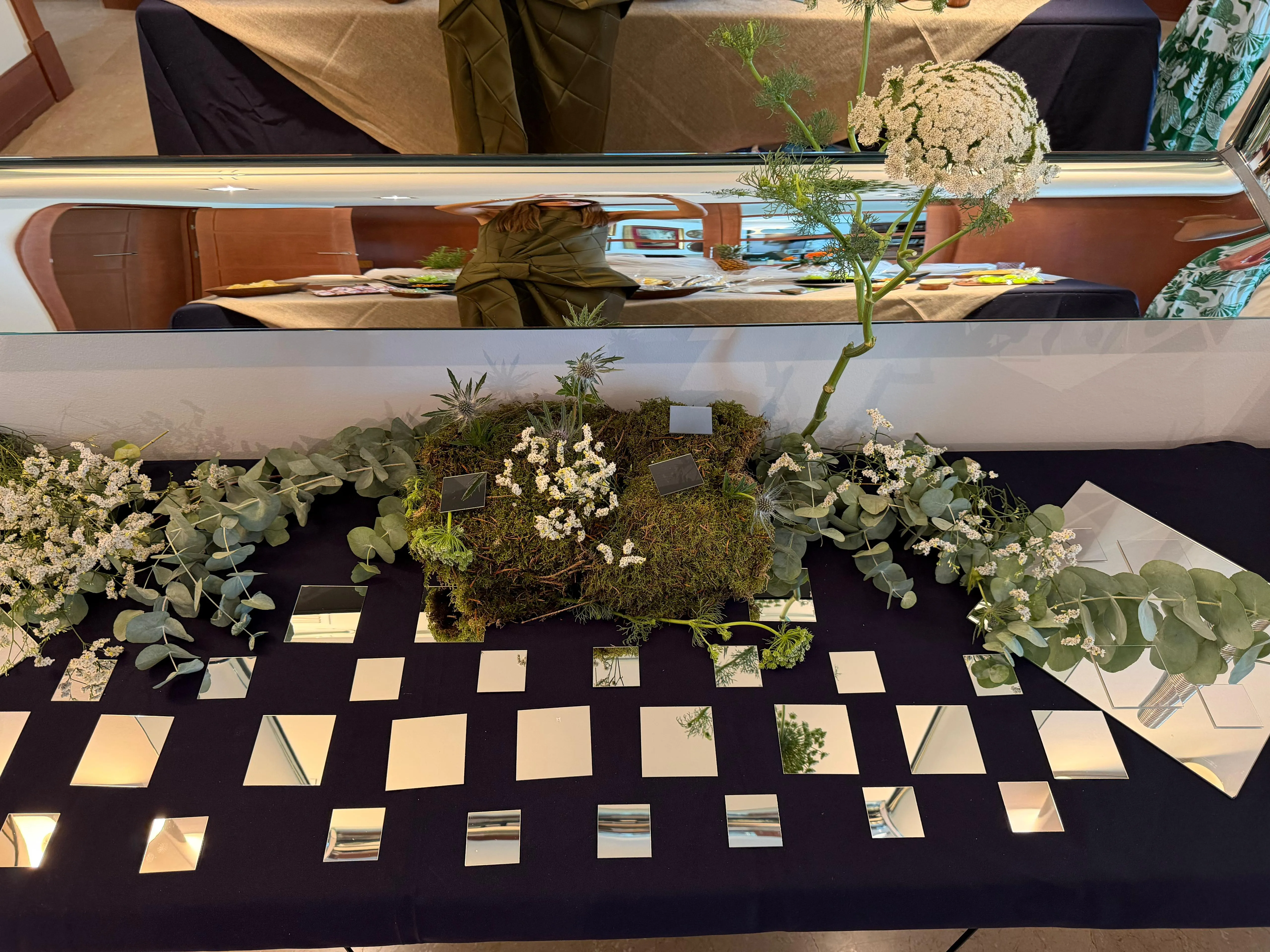 La mesa vacía antes del servicio: un lecho de musgo con flores blancas y eucalipto sobre mantel oscuro, rodeado de espejos cuadrados.
