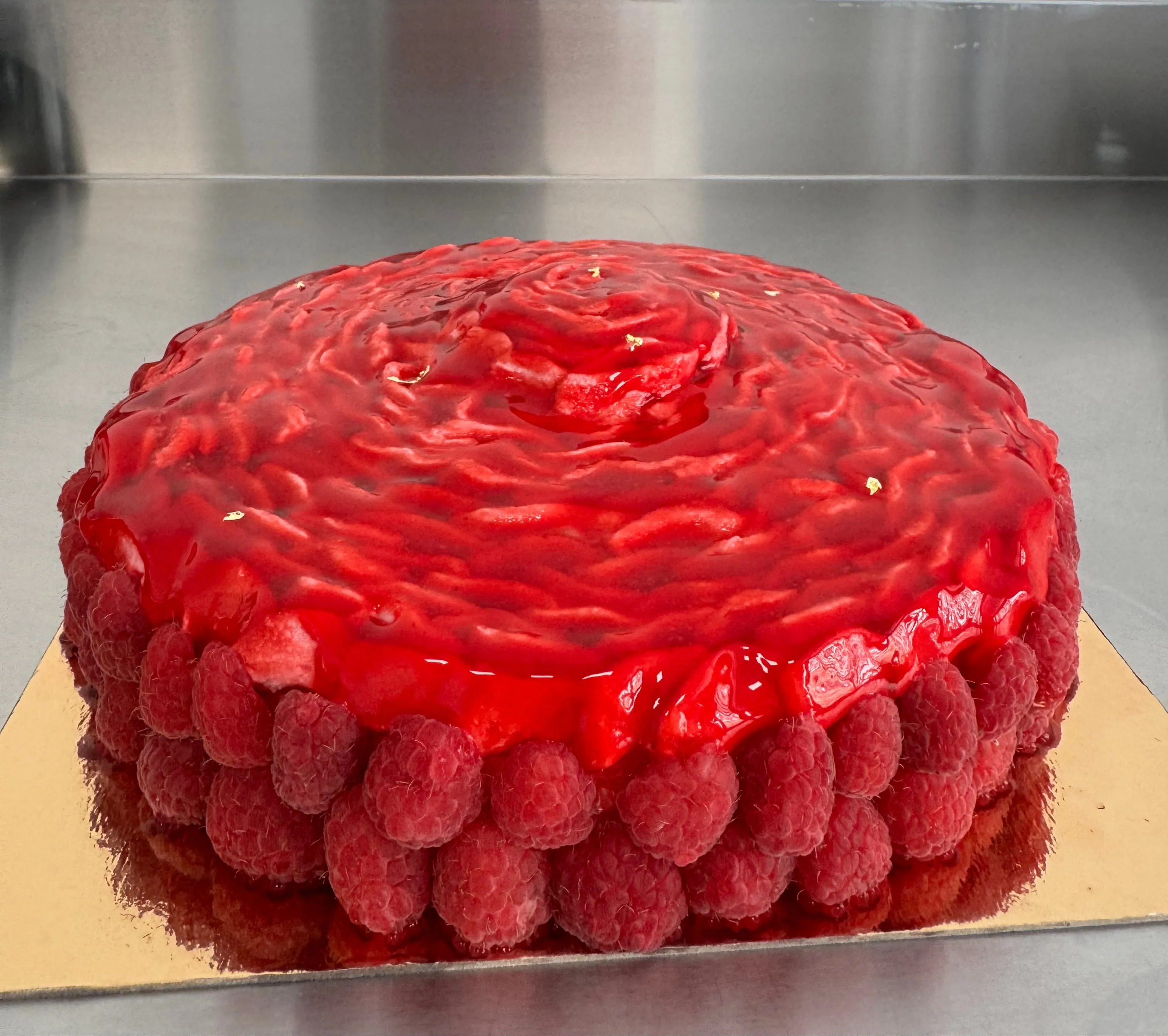 Tarta de glaseado rojo en espiral con un cerco de frambuesas, sobre una base dorada.